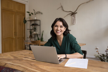 Joyful businesswoman sit at workplace with laptop at home office, smile look at camera feel...