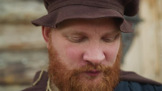 Red-bearded man in medieval clothes drinks potion from lid. Satisfied village squire uses magical elixir to sustain strength closeup against log house