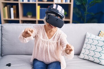 Middle age grey-haired woman playing motorbike video game using virtual reality glasses at home