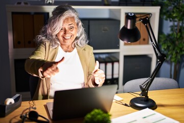 Middle age woman with grey hair working using computer laptop late at night approving doing positive gesture with hand, thumbs up smiling and happy for success. winner gesture.