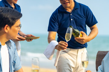 Happy Asian family couple and aging parents having celebration dinner during travel ocean together on summer holiday vacation. Waiter and waitress serving champagne to customers at beach restaurant.