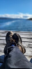 Vertical shot of the feet of a human with shoes sitting on the dock with the background of a sea