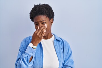 African american woman standing over blue background smelling something stinky and disgusting, intolerable smell, holding breath with fingers on nose. bad smell
