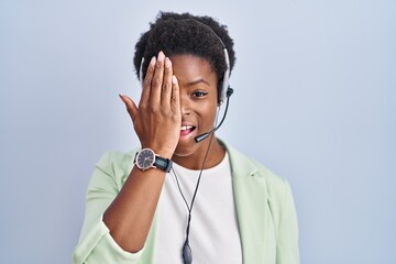 African american woman wearing call center agent headset covering one eye with hand, confident smile on face and surprise emotion.