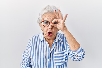 Senior woman with grey hair standing over white background doing ok gesture shocked with surprised...