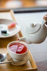 Image of a hand filling the cup with a pink tea from the white, small kettle.