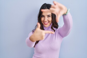 Young brunette woman standing over blue background smiling making frame with hands and fingers with happy face. creativity and photography concept.