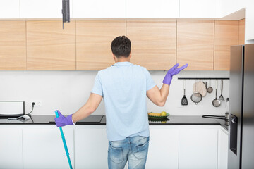 Back view on young man stands in the kitchen in rubber gloves, holds mop and doesn't know what to do