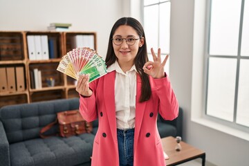 Young latin woman working at consultation office holding money doing ok sign with fingers, smiling friendly gesturing excellent symbol