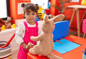 Adorable hispanic girl student looking for book standing backwards at kindergarten