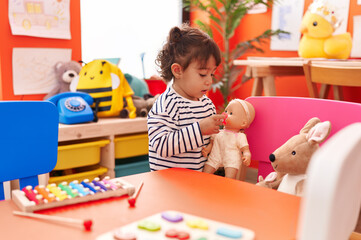 Adorable hispanic girl playing with baby doll standing at kindergarten
