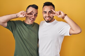 Homosexual couple standing over yellow background doing peace symbol with fingers over face, smiling cheerful showing victory