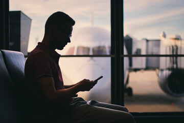Traveler using phone while waiting for flight. Silhouette of man waiting by window of airport terminal behind which is airplane..