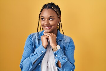 African american woman with braids standing over yellow background laughing nervous and excited...