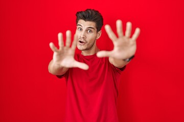 Young hispanic man standing over red background doing stop gesture with hands palms, angry and frustration expression