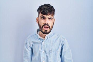 Obraz premium Young hispanic man with beard standing over blue background in shock face, looking skeptical and sarcastic, surprised with open mouth