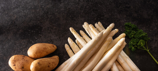 Fresh white asparagus and raw potatos on a modern black flagstone. Seasonal vegetables for preparing a healthy asparagus menu. Sunny kitchen scene for the spring gastronomy.