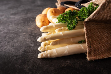 Fresh white asparagus and raw potatos in a shopping bag. Seasonal spring vegetables on modern black...