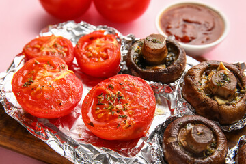 Aluminium foil with tasty baked mushrooms and tomatoes on pink background, closeup