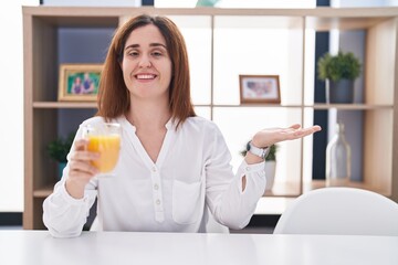 Brunette woman drinking glass of orange juice smiling cheerful presenting and pointing with palm of hand looking at the camera.