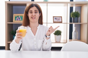 Brunette woman drinking glass of orange juice pointing aside worried and nervous with forefinger, concerned and surprised expression