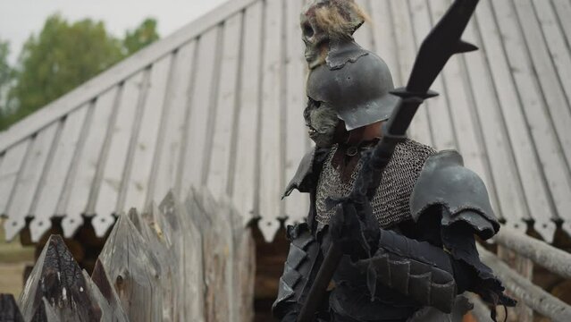 Knight in dark armor walks heavily along fence holding spear