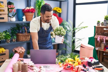 Young hispanic man florist using laptop holding plant pot at florist