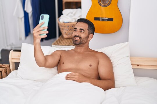 Young hispanic man make selfie by smartphone lying on bed at bedroom