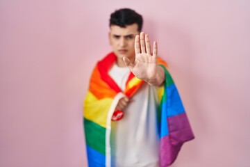 Non binary person holding rainbow lgbtq flag with open hand doing stop sign with serious and confident expression, defense gesture