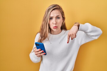 Young blonde woman using smartphone typing message pointing down looking sad and upset, indicating direction with fingers, unhappy and depressed.