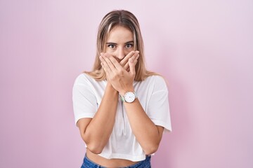 Young blonde woman standing over pink background shocked covering mouth with hands for mistake....