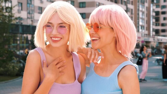 Two young beautiful smiling hipster female in trendy summer clothes. Sexy carefree women posing in the street in wigs. Positive funny models having fun at sunset. In sunglasses. At sunny day