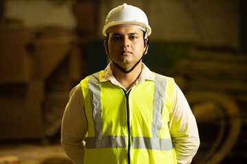 Portrait of Indian man engineer wearing safety hard hat and vest holding paper and pad standing in industrial factory. Closeup