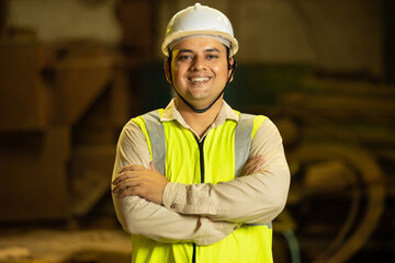 Portrait of happy Indian man engineer wearing safety hard hat and vest holding paper and pad standing with cross arms in industrial factory.