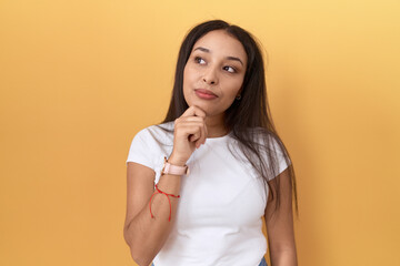 Young arab woman wearing casual white t shirt over yellow background with hand on chin thinking about question, pensive expression. smiling with thoughtful face. doubt concept.