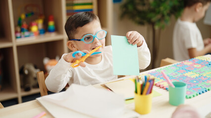 Adorable hispanic boy student smiling confident cutting paper at kindergarten