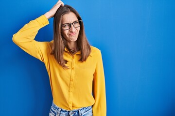 Young woman wearing glasses standing over blue background confuse and wonder about question....