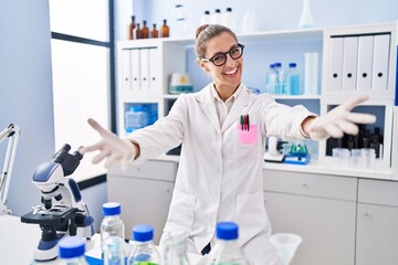 Young woman working at scientist laboratory looking at the camera smiling with open arms for hug....
