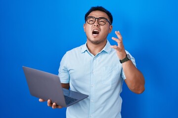 Chinese young man using computer laptop crazy and mad shouting and yelling with aggressive...