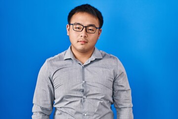 Young chinese man standing over blue background relaxed with serious expression on face. simple and natural looking at the camera.