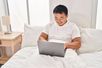 Young chinese man using laptop sitting on bed at bedroom