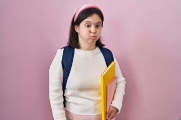 Woman with down syndrome wearing student backpack and holding books puffing cheeks with funny face. mouth inflated with air, crazy expression.