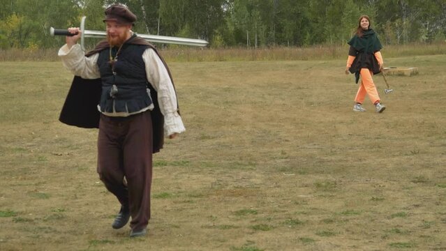 Red-bearded squire with sword and teenage girl with ax walk across lawn talking cheerfully. Medieval people prepare for combat training in nature horizon tilts
