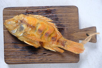 kan Nila Goreng is fried tilapia fish on a brown teak wood cutting board, on a white marble table. Tilapia is highly nutritious and a source of animal protein. Indonesian food