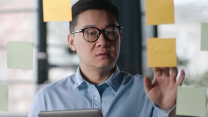 Serious middle aged asian man checking schedule in digital tablet, reading notes on stickers, standing near glass wall