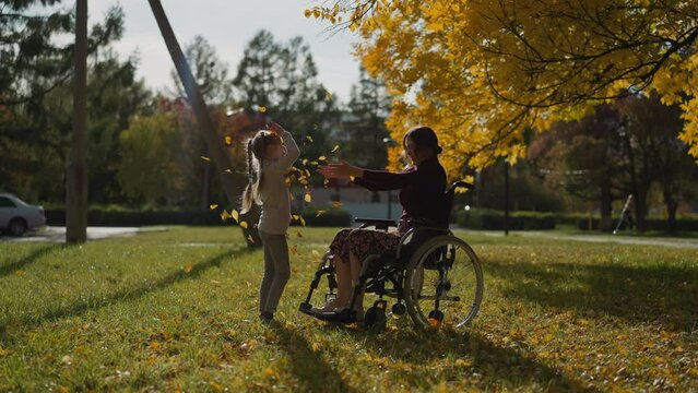 Woman with paralysis plays with daughter in park. Woman throws up dry leaves having fun with little girl. Active rest during rehabilitation process