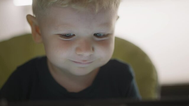 Happy little boy looks at glowing screen while rests in high chair at home closeup. Cute face of toddler child spending time on internet. Carefree childhood