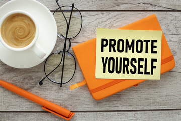 Promote Yourself . text on yellow sticky note and desk with orange notepad.