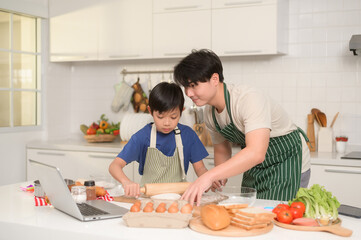 Happy young Asian father and kid are using a laptop computer to learn how to cooking food in their home kitchen.