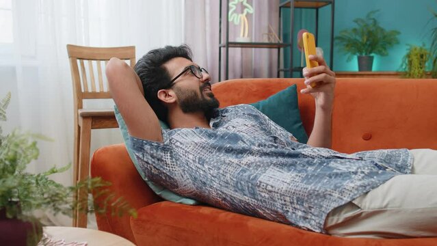 Indian man lying on couch uses mobile phone smiles at home living room apartment. Hindu young guy texting share messages content on smartphone social media applications online, watching relax movie